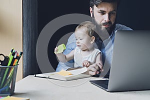 Baby sitting on knees of working parent. Father doing work at home sending projects online. Dad looking for vacancies on internet