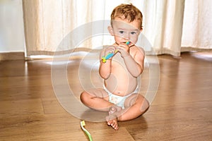 Baby sitting on a wooden floor of his house nibbling a toothbrush, while learning to brush his teeth