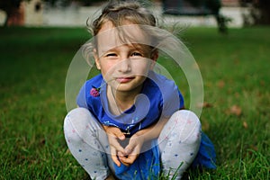 The baby is squatting and looking at the camera. The girl`s eyes narrowed ironically. She holds a purple clover flower