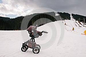 Baby stroller is on snow in the Carpathian mountains, outdoors. On background of the forest and ski slopes with lifts. Close up.