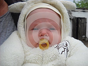 A baby sucks a weird rubber pacifier. Unusual, old-fashioned form of a baby`s dummy. The kid is dressed in a white fluffy jumpsui