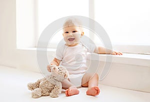 Baby with teddy bear toy sitting home in white room near wind