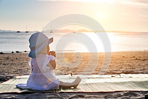 The baby is teething. Everything gnaws, chewing. Infant girl sitting on the tropical sand beach and looking for sunset