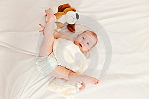 Baby less than a year lies on the bed with a soft toy. Baby in diaper lifts legs