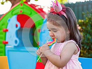 Baby toddler girl eating a large colorful lollipop dressed in pink dress as princess or queen