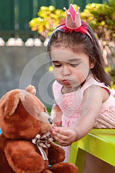 Baby toddler girl, mouth full of candies playing in a tea party feeds best friend Teddy Bear with candy