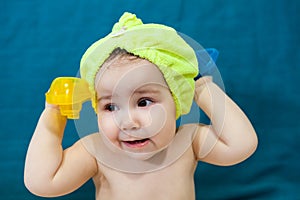 Baby with towel cap on head holding ship toys in hands, portrait after bath, blue background