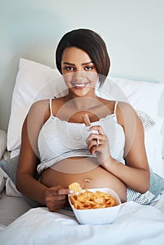 The baby wants what it wants. Portrait of an attractive young pregnant woman eating potato chips and chocolate in bed.