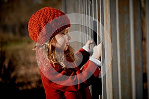 Baby 3 years with long hair.In a red beret and coat stands on the street in the sunshine, near the fence