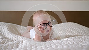 Babyhood, portrait of cute little child boy with big blue eyes in glasses lies on bed close up