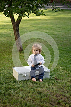 Babyin suit and necktie tie in green grass. Child having fun on family picnic in summer garden. Cute baby with suitcase