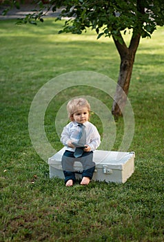 Babyin suit and necktie tie in green grass. Child having fun on family picnic in summer garden. Cute baby with suitcase