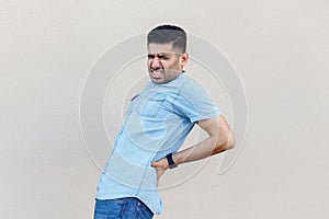 Back ache or kidney pain. Portrait of sick handsome young bearded man in blue shirt standing and holding his painful back