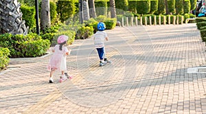 Back Asian little kid boy and girl wear safe helmet play kick board on road in park outdoors on summer day