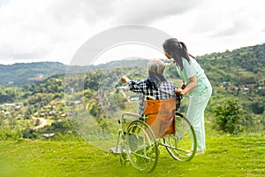 Back of Asian nurse stand and look to the direction of senior man on wheelchair pointing and both stay near cliff with village and