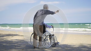 Back elderly woman disabled sitting in wheelchair and husband is a wheelchair user on the beach