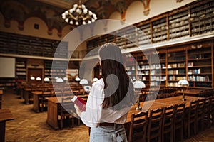 Back of female student in university campus library with book in hand. Studying in library