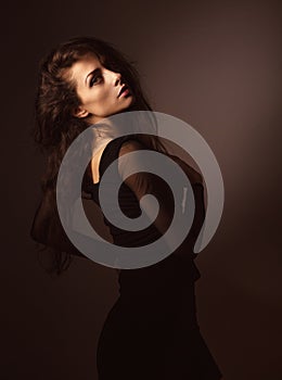 Back female view. Beautiful elegant sensual woman with brown long hair and black dress looking on dark shadow background.