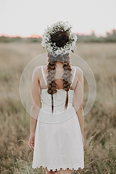 Back of the girl in a white dress in boho style with braids and a floral wreath in the summer in the field
