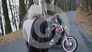 Back look of blonde comes to her smiling biker sitting at motorcycle in park