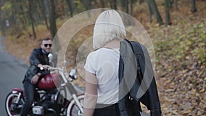 Back look of blonde comes to her smiling brutal biker with motorcycle in park