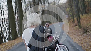 Back look of blonde in love comes to her biker sitting at motorcycle in park