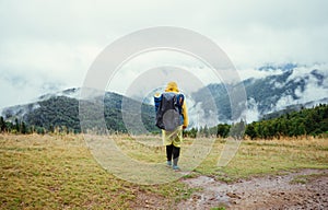 The back of a male tourist in a yellow raincoat walks on a meadow in the mountains against the backdrop of mountain views with