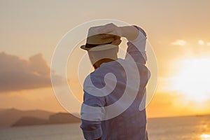 The back of a man in a hat against the background of the sea in the bright golden rays of the sun at sunrise