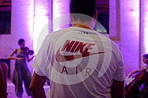 Back of a man in a nike shirt. In the background a girl plays the violin in the night lights