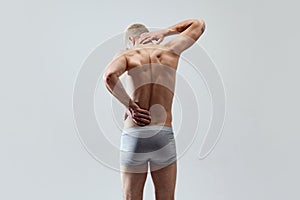 Back pains. Rear view image of young man with muscular, relief, body, back, posing in underwear shirtless against grey