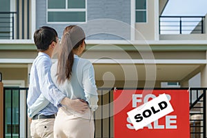 Back portrait of Asian young couple standing and hugging together looking happy in front of their new house to start new life.