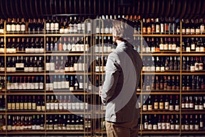 Sommelier standing in wine cellar, shelf with many bottles background and looks for necessary alcoholic beverage, make a photo