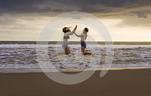 Back portrait of two happy and attractive young women girlfriends holding hands on the beach running to the sea under beautiful su