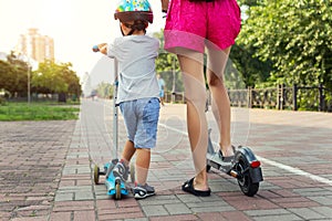 Back rear view of cute little baby boy with young adult mother riding scooter transport on city street park together on