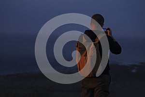 back rear view of one alone backpacker Man with rucksack, cell phone standing on peck of rock, taking photos of the view. Man on