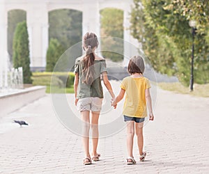 Back rear view two little girls 7 and 9 years old wearing casual clothes going on park green sunshine lawn