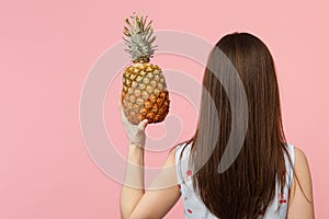 Back rear view of young woman in summer clothes holding in hand fresh ripe pineapple fruit isolated on pink pastel wall