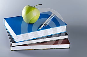 Three books with an apple, pen and pencil on grey background.