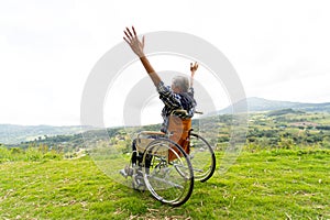 Back of senior man sit on wheelchair and action of raise his hands up like stretching also stay near cliff with mountain and grass