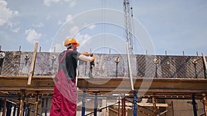 Back shot of professional construction worker in hard hat and bib overall makes formwork with electric screwdriver for