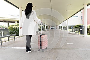 Back shot of woman in sneakers and coat walking towards main entrance of airport and rolling her wheeled suitcase.