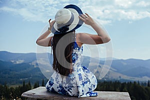 Back shot of young woman in elegant dress admiring breathtaking view while sitting on bench in the mountains