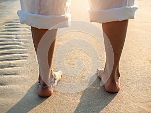 Back side of barefoot woman in white pants with folded legs standing on clean sandy beach in the warm sunlight.