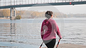 Back side follow view of young chubby girl doing morning nordic walking exercise alone river and city park