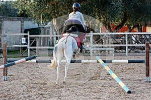 Back side of Girl riding a white horse jumping a guided obstacle during equestrian school training