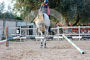 Back side of Girl riding a white horse jumping a guided obstacle during equestrian school training