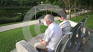 Back and side view of elderly retired Asian couple wearing face mask, sitting on bench.