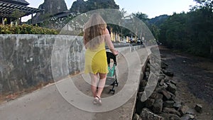 Back side view. A young mother in a yellow dress walks with the infant baby girl in a stroller along the promenade of a tropical r