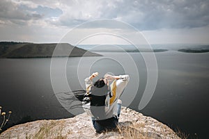 Back of stylish hipster girl sitting on top of rock mountain with beautiful view on river. Young tourist woman with windy hair