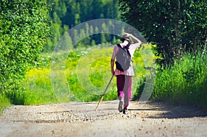 Back of a Teenager in a Pink T-Shirt, Red Pants, and Bucket Hat, with Black Backpack and Long Pole Walks along Mountain Country